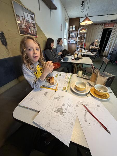 A cozy cafe scene unfolds at Commandantsbrug, Marineterrein, Amsterdam, where a child is engrossed in coloring while adults relax with knitting and drinks